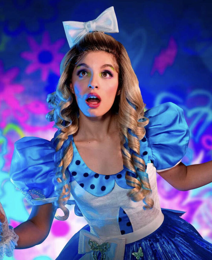 Femme en costume coloré et fantaisiste "Alice et la Reine de Coeur" avec une expression surprise sur un fond de type graffiti.