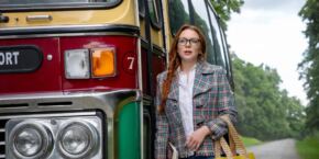 Femme debout à côté d’un bus d’époque sur une route bordée d’arbres, ressemblant à une scène tout droit sortie d’un film Marvel.