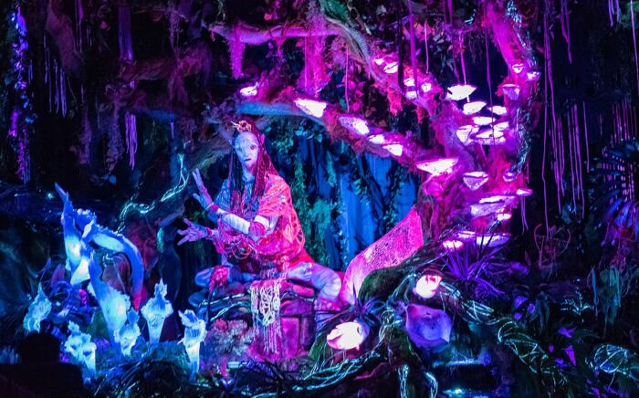 Une scène colorée et mystique dans Pandora – Le monde d'Avatar, avec une personne vêtue d'une tenue élaborée, entourée de champignons lumineux et de plantes fluorescentes au Walt Disney World Resort.