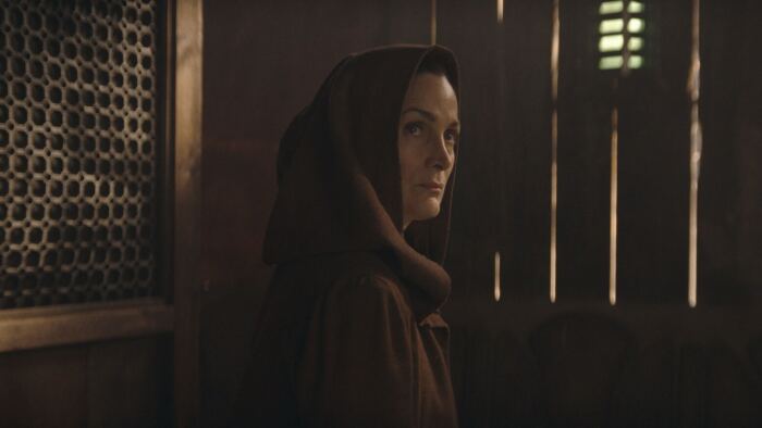 Une femme vêtue d'une robe à capuche, identifiable comme un Acolyte, debout dans une pièce faiblement éclairée avec des lumières verticales rappelant Star Wars, en arrière-plan.