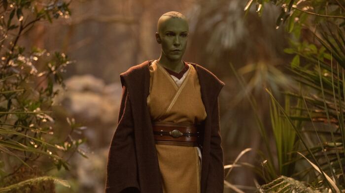 Une personne en costume de Jedi à la peau verte et à l'expression sérieuse se tient dans un décor aux allures de forêt, rappelant les scènes de The Acolyte sur Disney+.