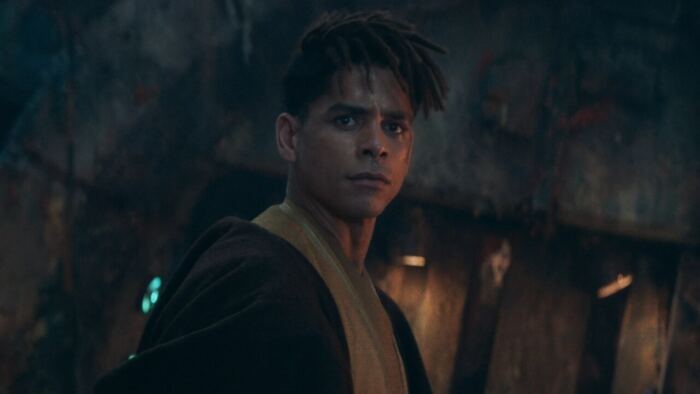 Un jeune Acolyte au regard intense se tenant dans un environnement faiblement éclairé avec un décor Star Wars en détresse.
