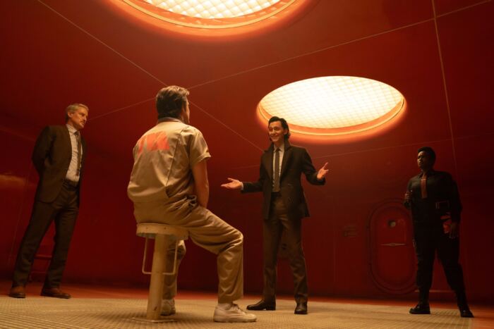 Quatre hommes conversant dans une pièce avec un éclairage rouge intense et un plafonnier circulaire, discutant de leur avis sur la série Loki saison 2.