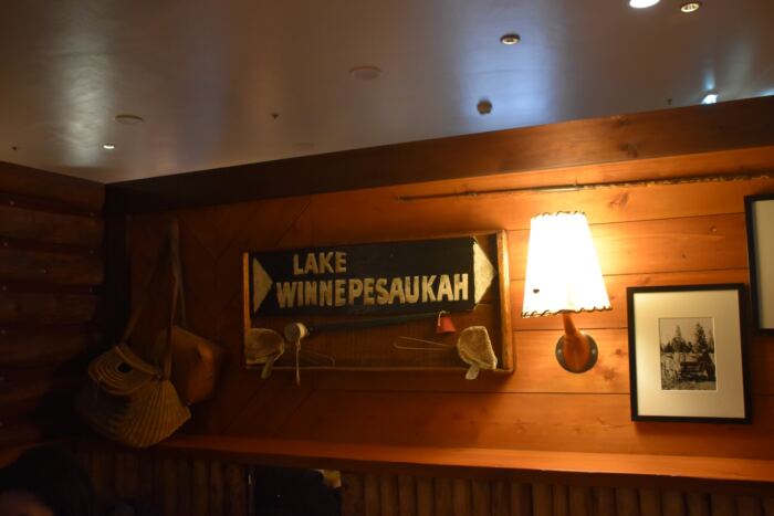 Un mur en bois comporte une pancarte indiquant « Lac Winnepesaukah » et est orné d'une canne à pêche, d'un panier et d'une applique murale allumée. Une photographie encadrée est accrochée sur le côté droit.