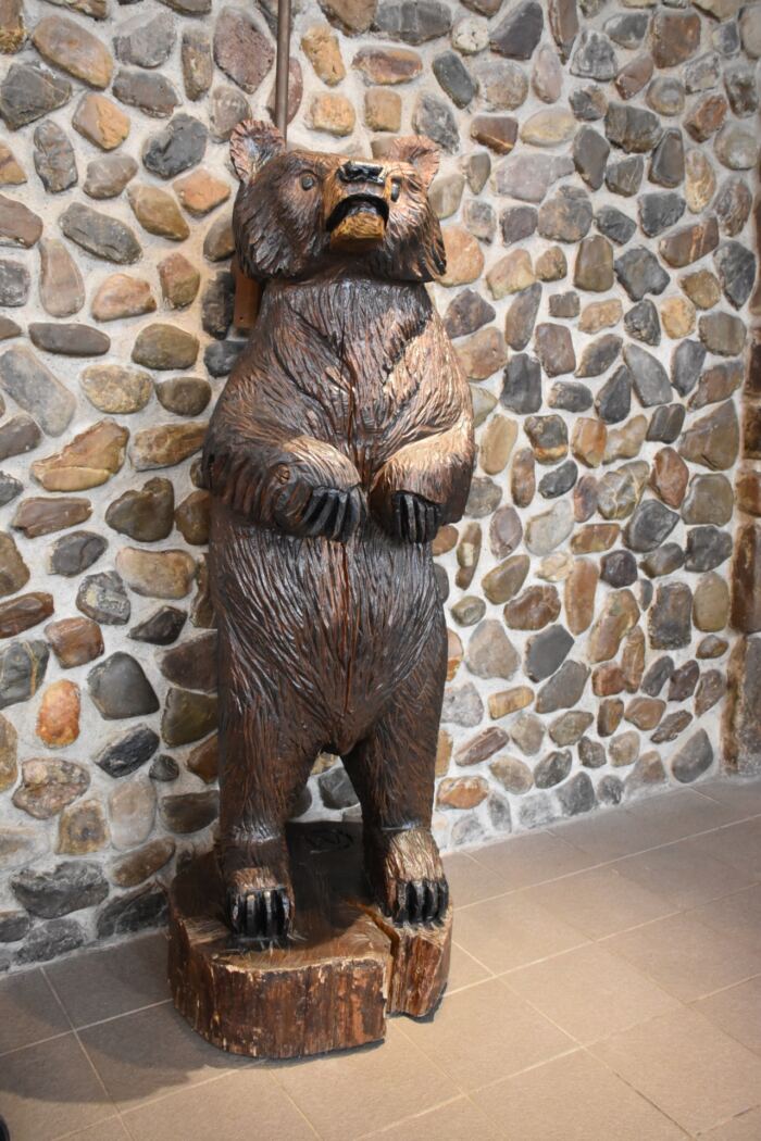 Une sculpture d'ours en bois se dresse sur un sol à motifs de pierre, sur fond de mur pavé. L'ours est debout, les pattes avant légèrement relevées.