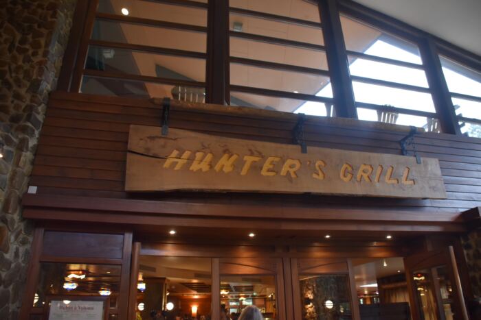 Panneau en bois indiquant "Hunter's Grill" au-dessus de l'entrée d'un restaurant doté de grandes fenêtres et d'un éclairage intérieur chaleureux.