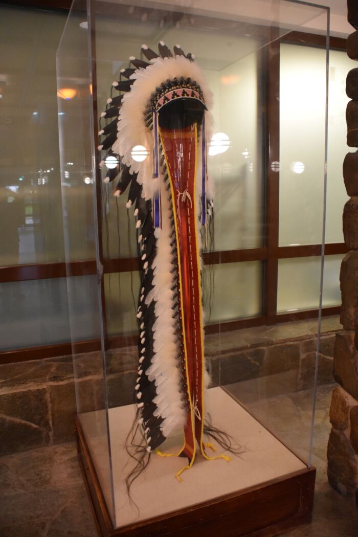 Une coiffe en plumes amérindienne exposée dans une vitrine avec un fond comportant un mur et des fenêtres en pierre.