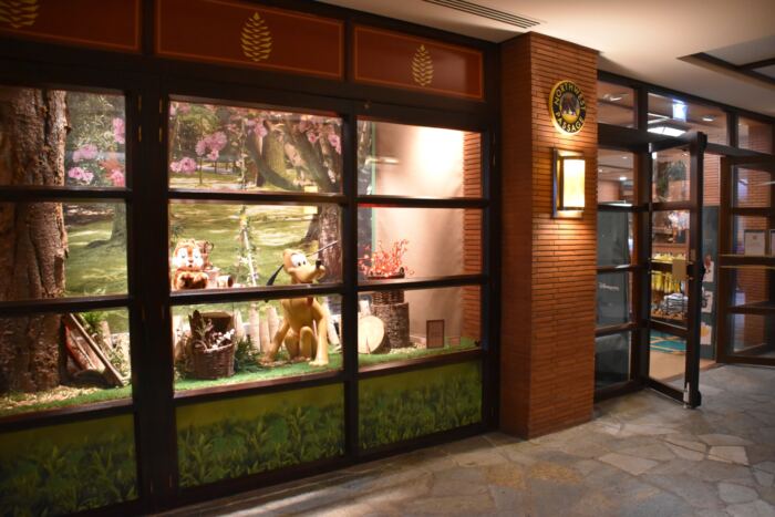 Présentoir de vitrine présentant deux statues de cerfs, un panier et de la verdure devant un décor sur le thème de la nature. La porte vitrée à droite montre l'intérieur du magasin avec des étagères de produits.