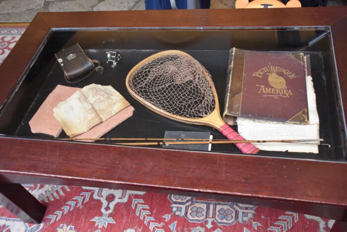 Une table en bois avec plateau en verre présente un filet de pêche à la mouche, un livre ancien intitulé « L'Amérique pittoresque », des cartes pliées, un petit étui en cuir et un étui à stylos avec des tiges attachées, posés sur un tapis à motifs.