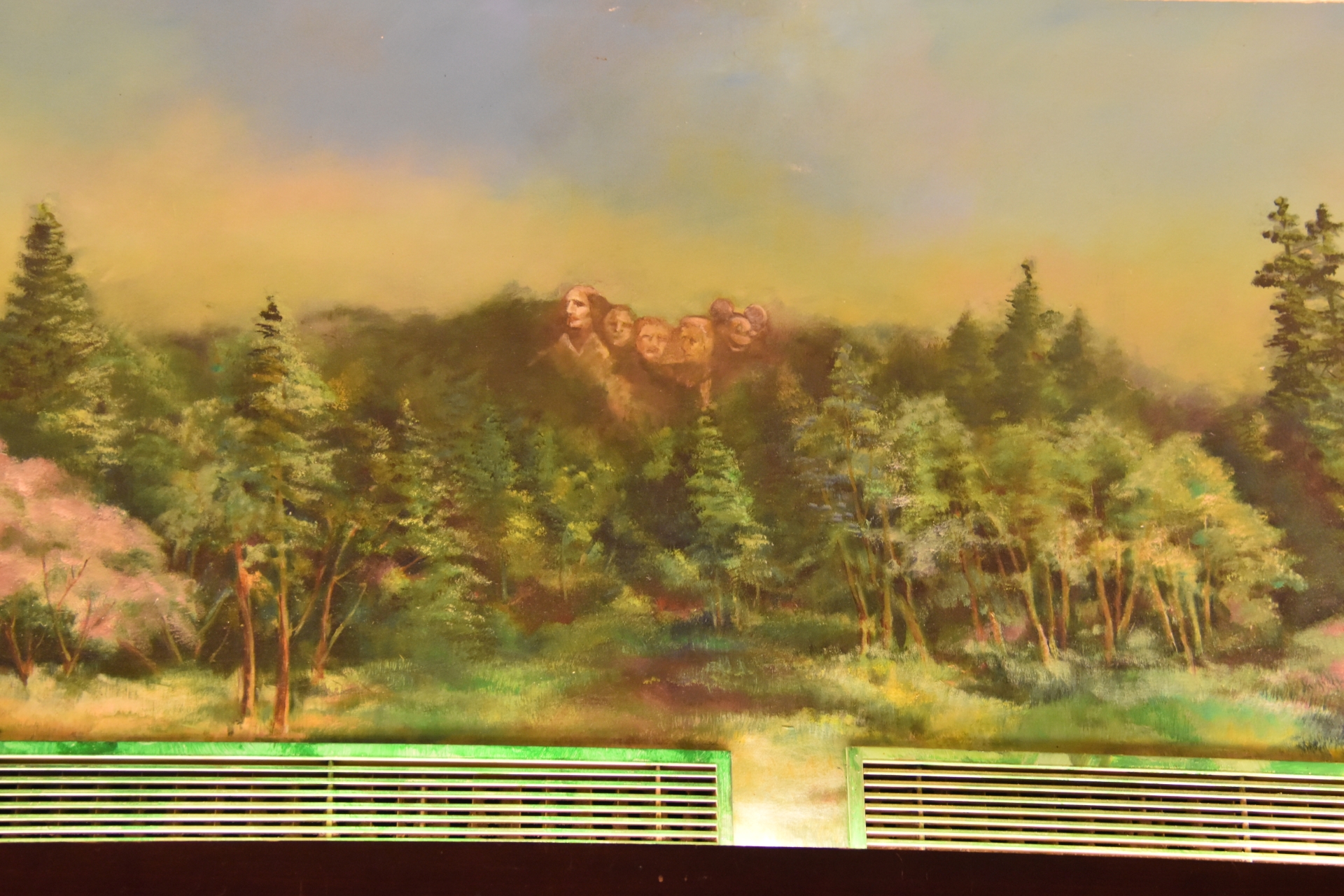 Un tableau représente le mont Rushmore entouré d'une forêt. Le ciel est d'une couleur brumeuse et jaunâtre et l'œuvre d'art est encadrée par des grilles de ventilation en bas.