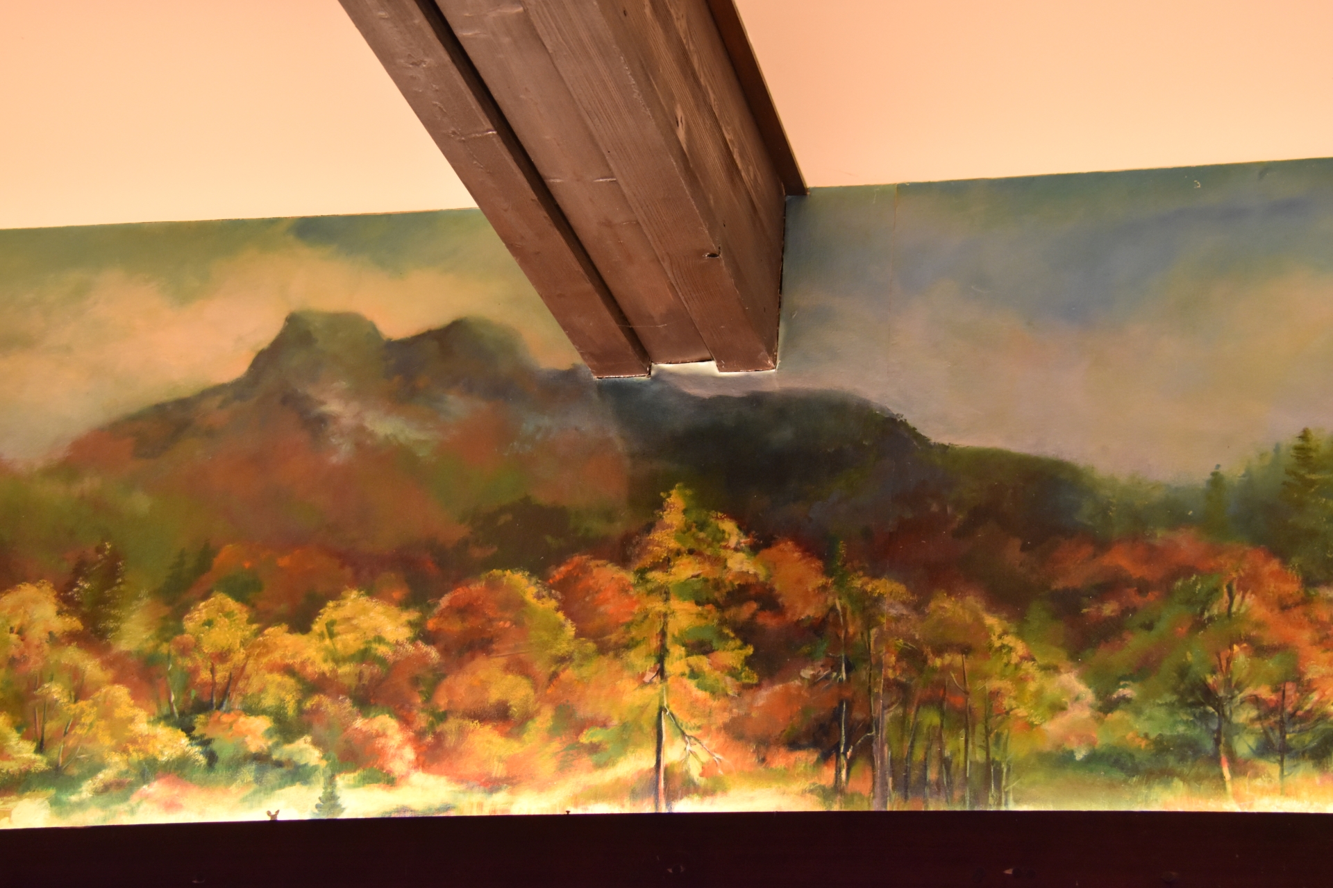 Une peinture murale représentant une forêt en automne avec un feuillage automnal coloré, avec une montagne en arrière-plan, partiellement recouverte par une poutre de plafond en bois.