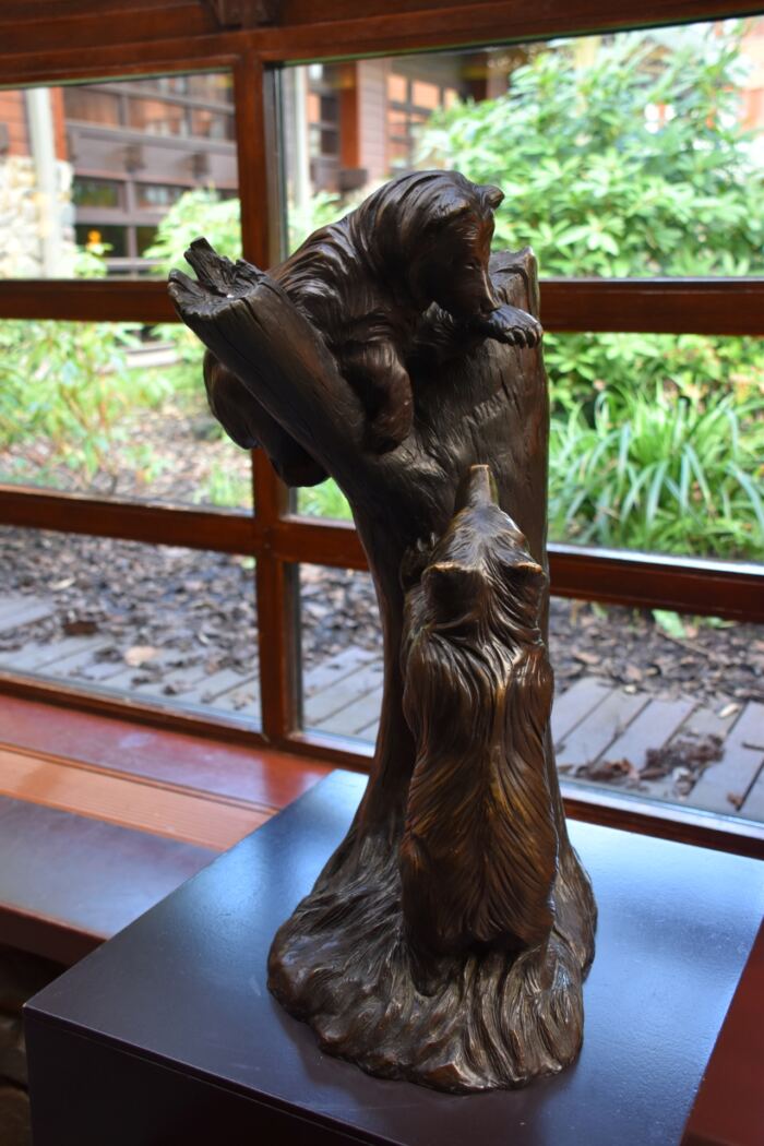 Une sculpture en bronze représentant deux ours en interaction, l'un grimpant sur une souche d'arbre tandis que l'autre lève les yeux, exposée sur un piédestal devant une fenêtre avec vue sur le jardin à l'extérieur.