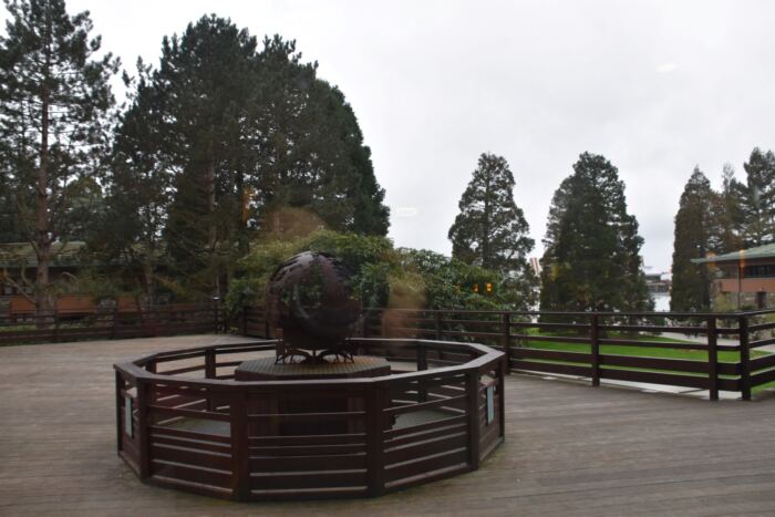 Une sculpture en forme de sphère métallique repose sur une terrasse en bois entourée de balustrades, avec des arbres et des bâtiments visibles en arrière-plan par temps nuageux.