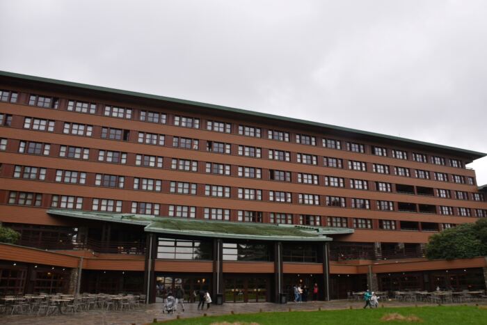 Un grand bâtiment à plusieurs étages avec un extérieur marron et de nombreuses fenêtres. Les gens sont rassemblés près de l’entrée par une journée nuageuse. Des tables et des chaises d'extérieur sont disposées à proximité.