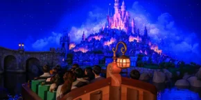 Une promenade en bateau avec des passagers assis s'approche d'un château fantastique illuminé sur fond de ciel nocturne bleu vif, rappelant les attractions enchanteresses des parcs Disney à l'étranger.