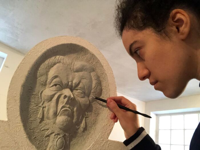 Dans une pièce bien éclairée rappelant un décor de cinéma, une personne sculpte méticuleusement un visage détaillé sur une surface en pierre, son outil de sculpture capturant l'essence étrange de Phantom Manor.