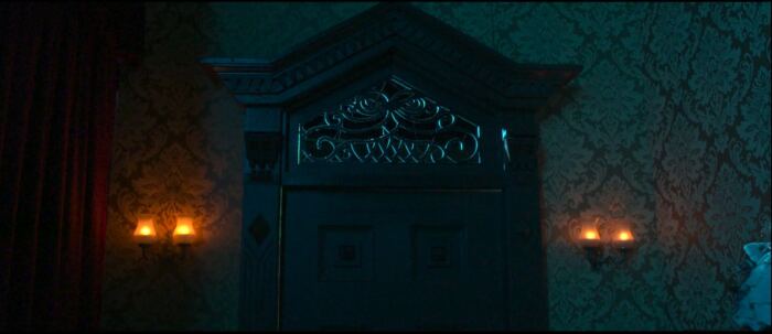 Une porte en bois ornée, faiblement éclairée, avec des sculptures complexes, rappelant l'attrait mystérieux de Phantom Manor, est encadrée par du papier peint à motifs et deux appliques murales avec des bougies allumées de chaque côté, comme si elle plantait le décor d'un moment du film.