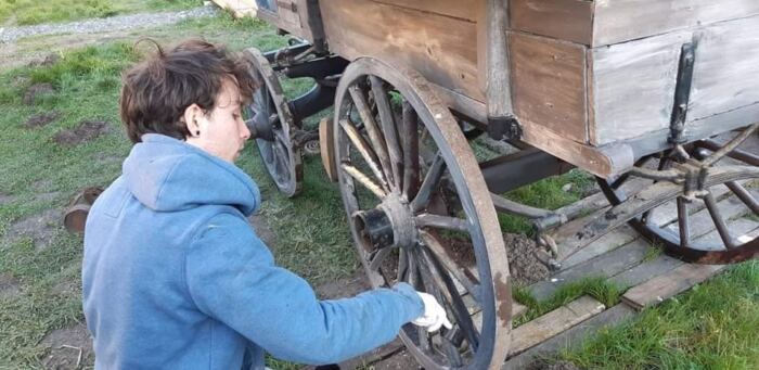 Une personne portant un sweat à capuche bleu répare la roue en bois d'un vieux chariot à l'extérieur sur une zone herbeuse, créant une scène rappelant un décor de film classique près de Phantom Manor.