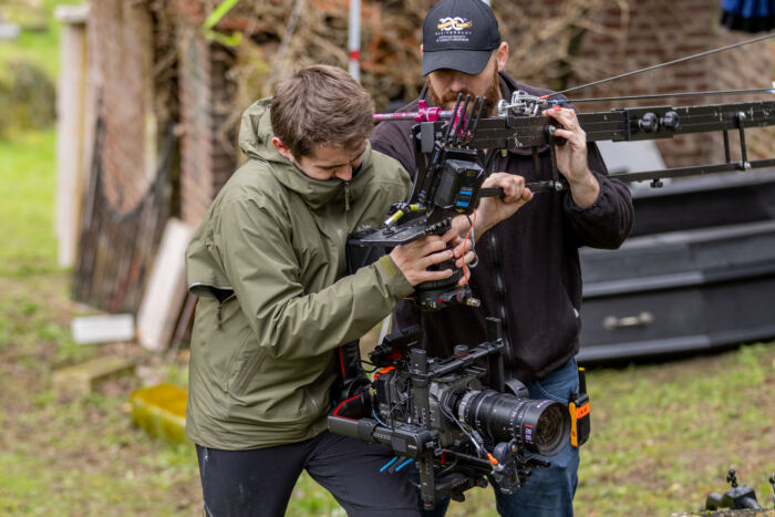Deux personnes ajustent un support de caméra en extérieur, comme des réalisateurs de Phantom Manor. L'un porte une veste verte, l'autre une casquette. Ils se tiennent côte à côte près de structures en brique et en bois, préparant leur prochaine prise de vue cinématographique.